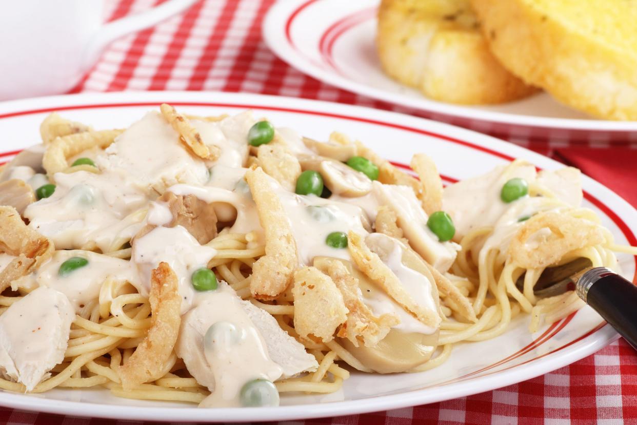 Chicken tetrazzini on a white plate with a red stripe and a fork on a red checkered tablecloth with a side of buttered toast