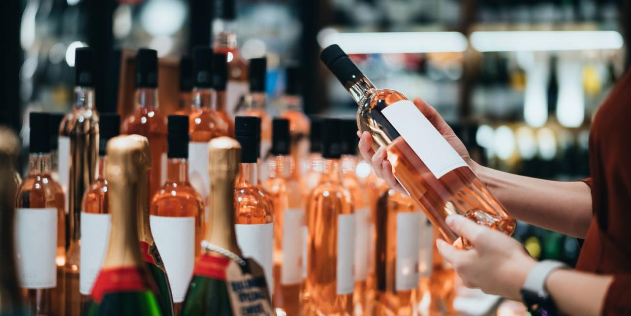 close up of young asian woman walking through liquor aisle and choosing a bottle of rose wine from the shelf in supermarket