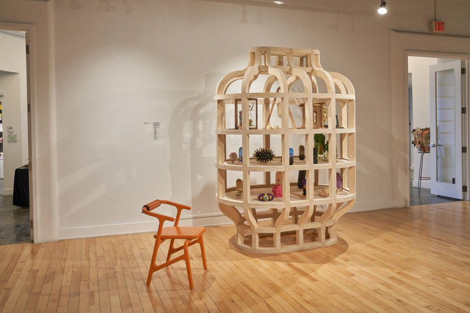 Norman Teague's Cabinet of Curiosities, features a shelf-like cabinet of wood with various small objects on the shelves, next to a structural orange chair.
