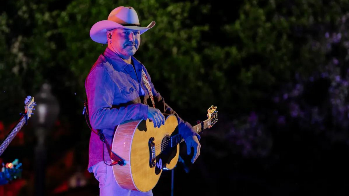 A white man looks towards the camera. He is wearing a white cowboy hat and holding a guitar. 