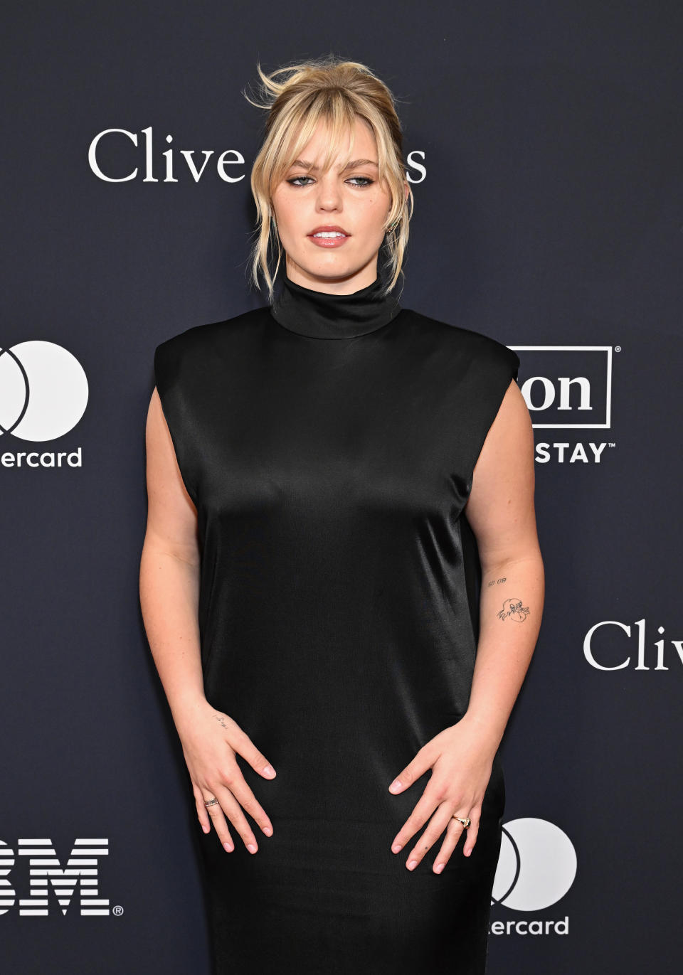 Reneé in a sleeveless turtleneck dress posing on red carpet; she wears minimal jewelry and has a tattoo on her arm