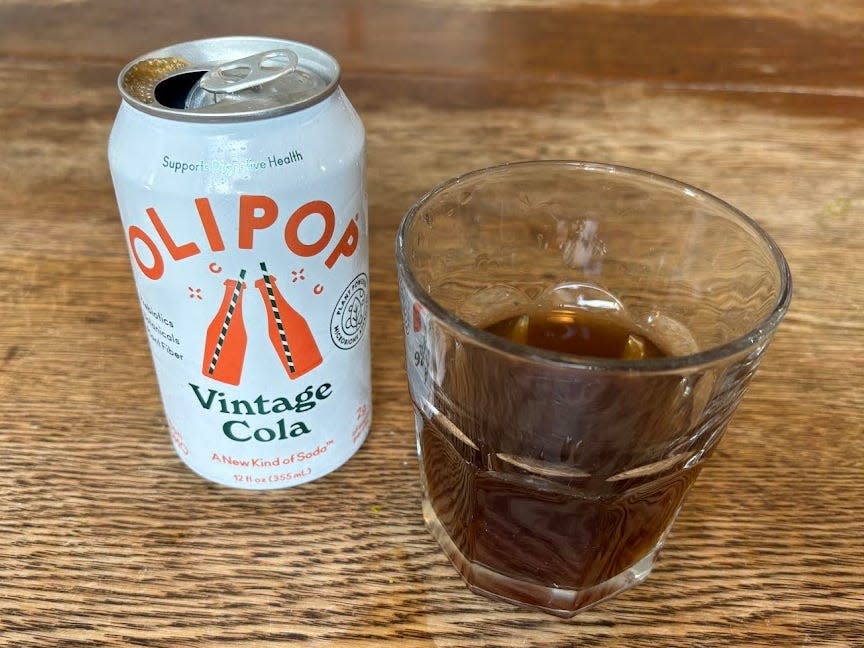 An open can of vintage cola Olipop next to a small, clear glass with brown liquid inside. Both are sitting on a wooden table.