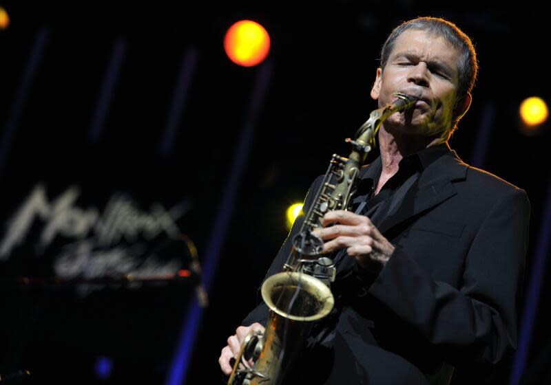 A man in a black blazer holding and playing a saxophone on a dark stage