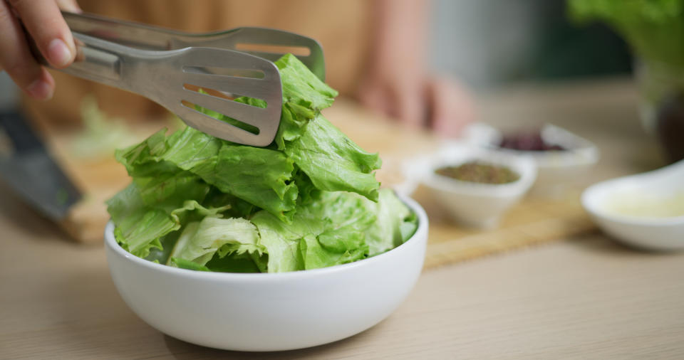 A person holds tongs, placing fresh lettuce leaves into a white bowl with small dishes of spices and herbs visible in the background