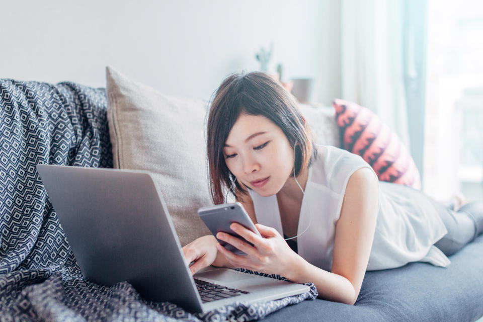 A woman is lying on a couch, using a laptop and holding a smartphone. She appears to be working from home