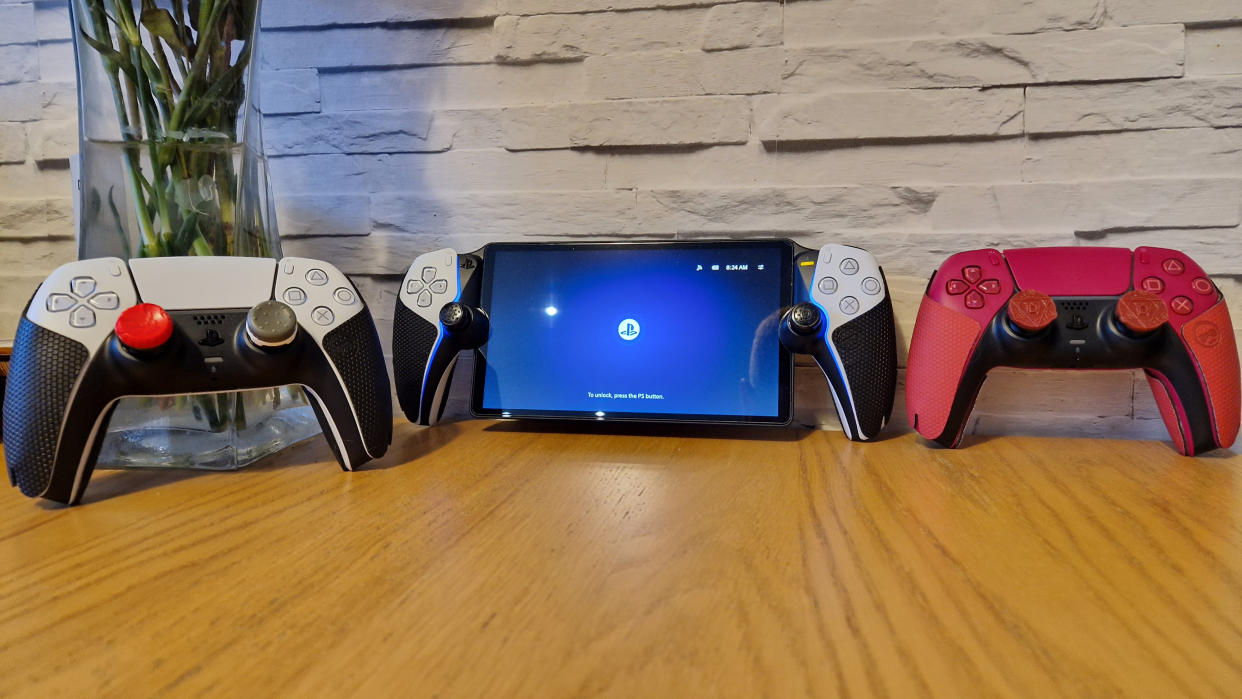  Two DualSense controllers and a PlayStation Portal with thumbsticks and grips attached on a wooden surface with a white brick background. 