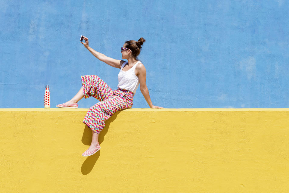 Junge modische Frau sitzt auf gelber Mauer und macht ein Selfie