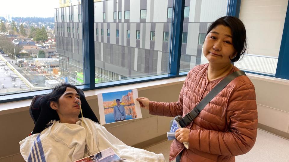 Zhenhuan Lei sits in a wheelchair at Royal Columbian Hospital in New Westminster as his mother, Ying Li, holds a photo of her son before his hospitalization. Li has filed a lawsuit alleging people and groups connected to an Oct. 2023 martial arts tournament are responsible for an injury that led to her son's vegetative state.