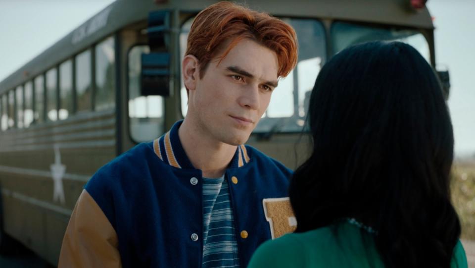 Archie Andrews wears a letterman jacket and stands before a bus talking to Veronica in a scene from Riverdale.