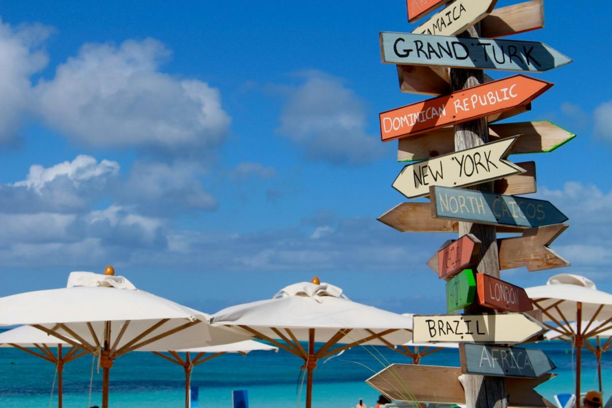Turks and Caicos is a great travel destination that is considered safe. Learn more about what travelers should know. pictured: a signpost with international destinations in front of beach umbrellas on a cloudy Turks and Caicos day
