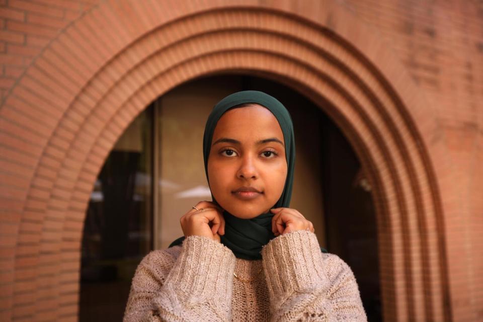 A woman in a head covering stands in front of a brick arches doorway outside a building.