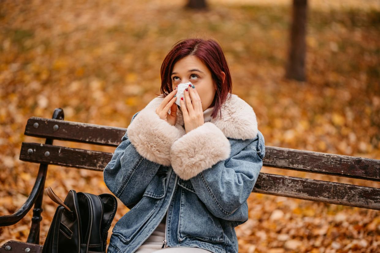 a young woman in a jacket sitting on a park bench and wiping her face with a tissue amidst fall foliage, rolling her eyes in exasperation at seasonal allergies.