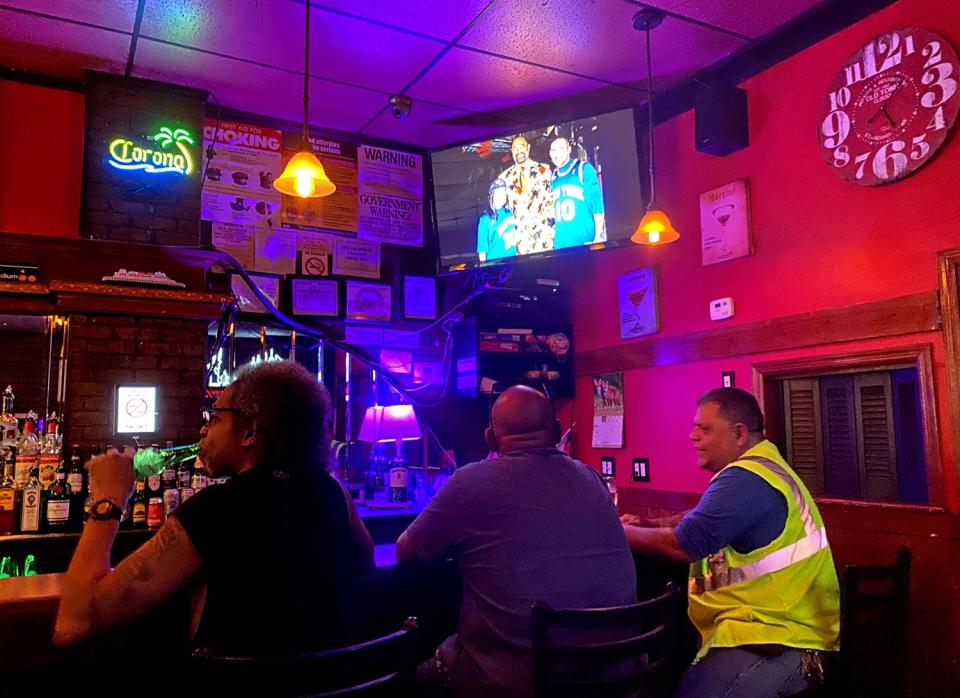 Men sit at a bar drinking, with a large TV hanging overhead.