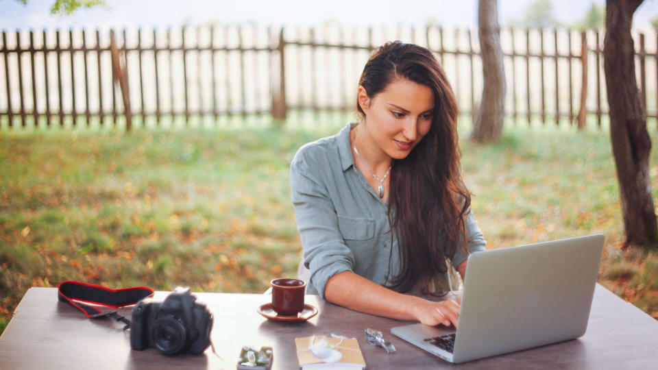 Smiling female novelist, blogger, freelancer or a photographer, typing on the laptop keyboard while having a cup of coffee, sitting in the bright outdoors at the desk.