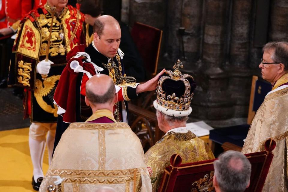 prince william stands before king charles who is seated and wearing a large crown, william touches the crown and looks down, both men wears robes as other men stand and watch