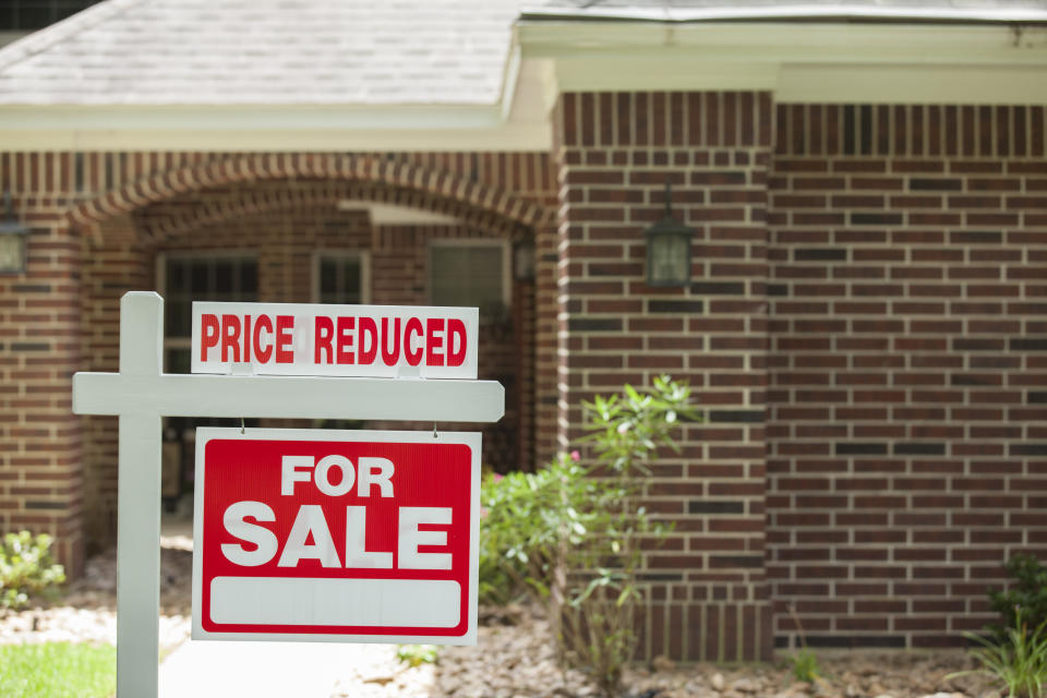 Red and white "Price Reduced, Home for Sale" sign in front of a red brick house that is for sale and its price has been reduced due to slow economy.  Green grass and bushes indicate the spring or summer season. Front porch and windows in background. Real estate sign in residential neighborhood.  Moving house, relocation concepts.