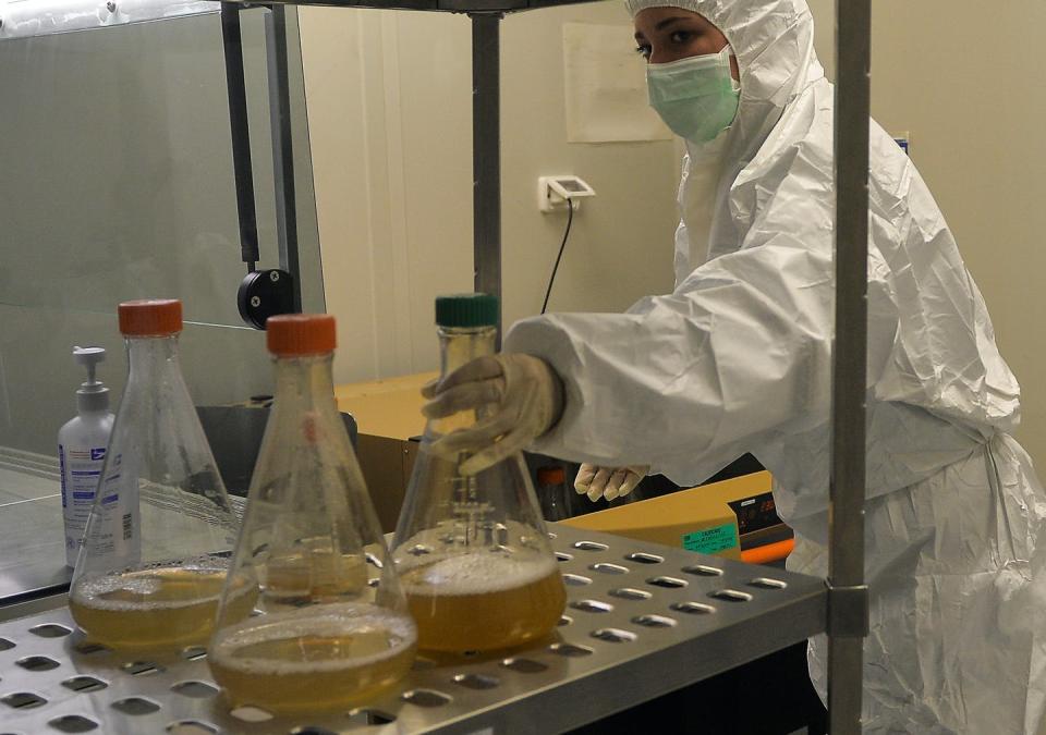 A woman in full body PPE reaches for a beaker containing a murky liquid.