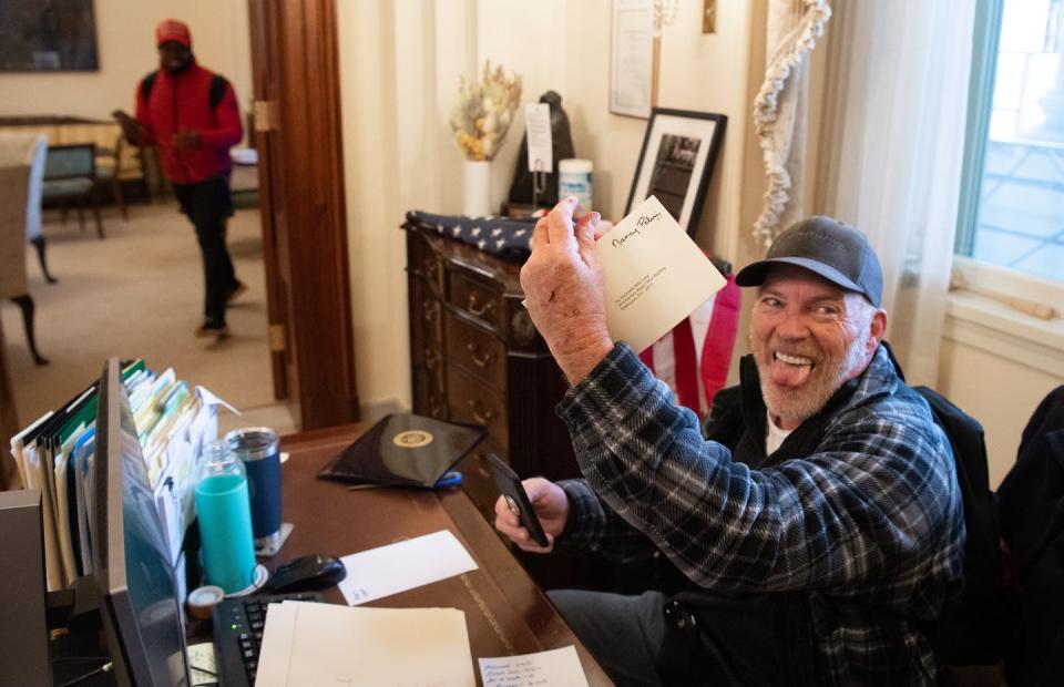 Richard Barnett sits at a desk holding a piece of mail with Nancy Pelosi's name and sticking his tongue out.