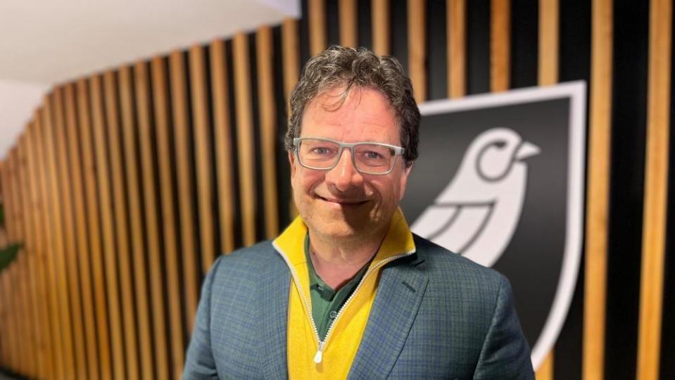 A smiling Mark Attanasio with brown hair and glasses in front of a Norwich City logo