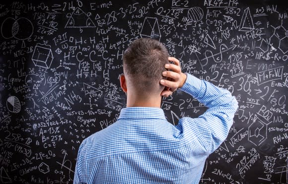 A man holding his hand to his head and facing a chalkboard on which are drawn mathematical formulas.