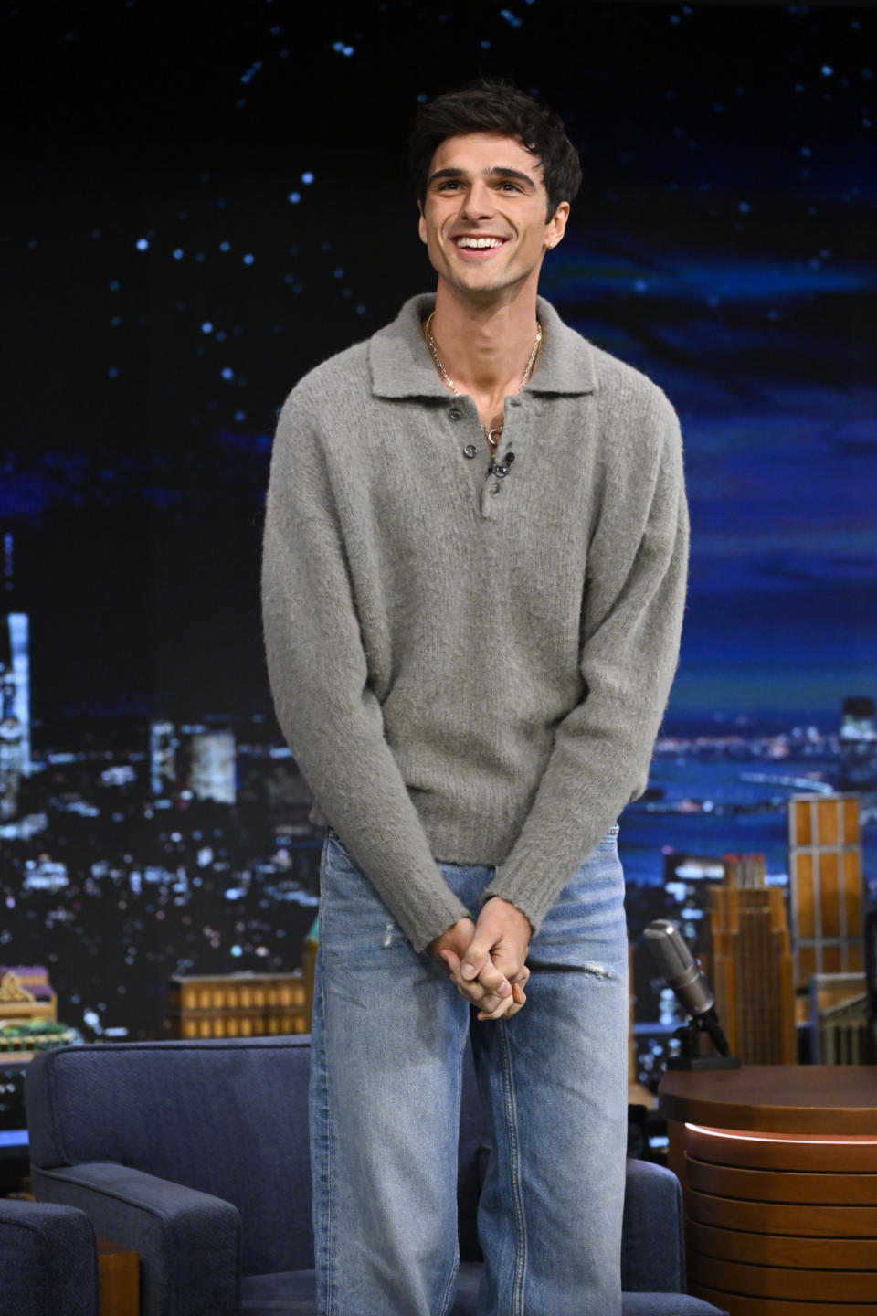 Jacob Elordi stands on a talk show set, smiling, wearing a casual grey sweater and jeans