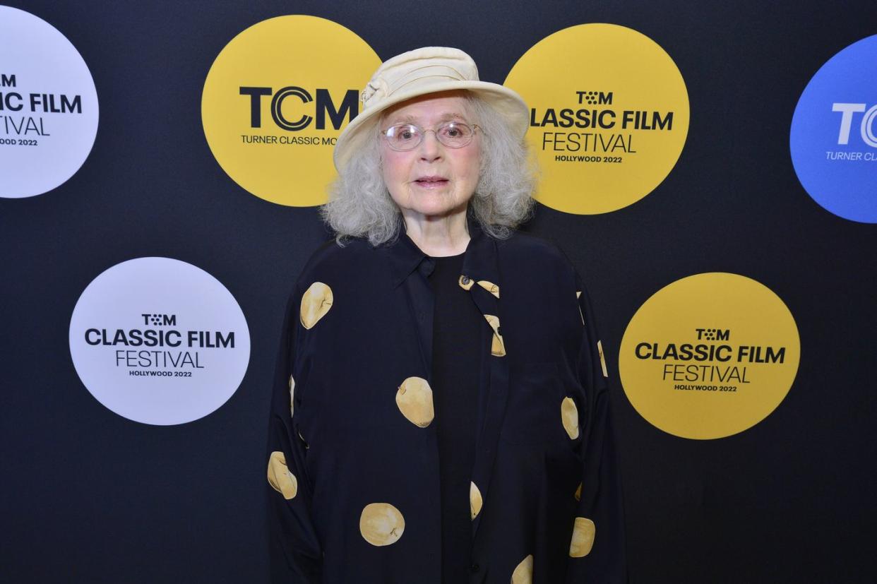 piper laurie wearing thin framed glasses, a white bucket hat and a black shirt with yellow polka dots, standing in front of a backdrop with various yellow and purple dots that read 'tcm classic film festival hollywood 2022'