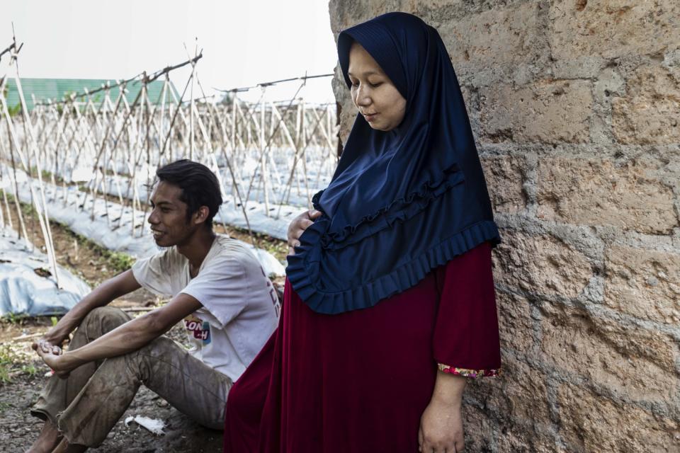 A woman in blue hijab and red robe stands with a hand on her stomach, next to a seated man who is smiling