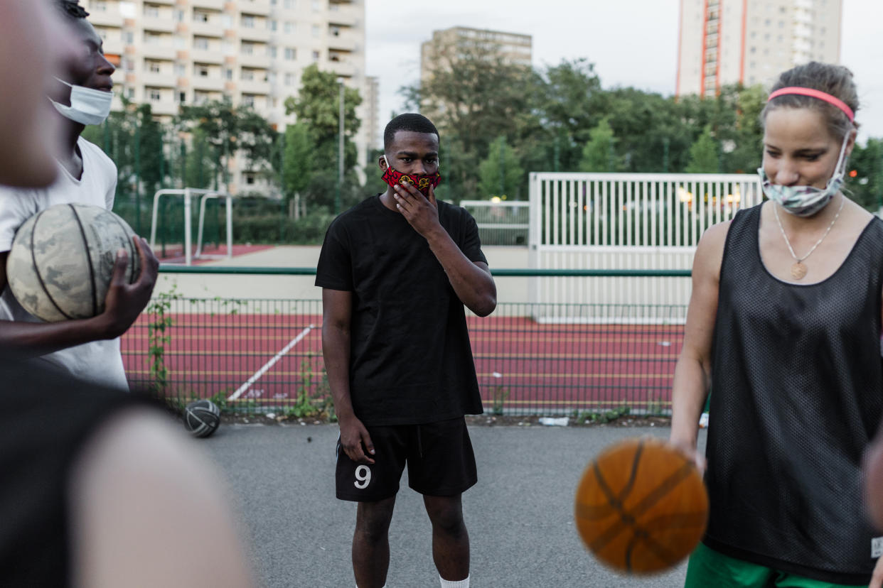 Black man wearing homemade facemask  He is standing at a distance from the other players on a Basketball court.