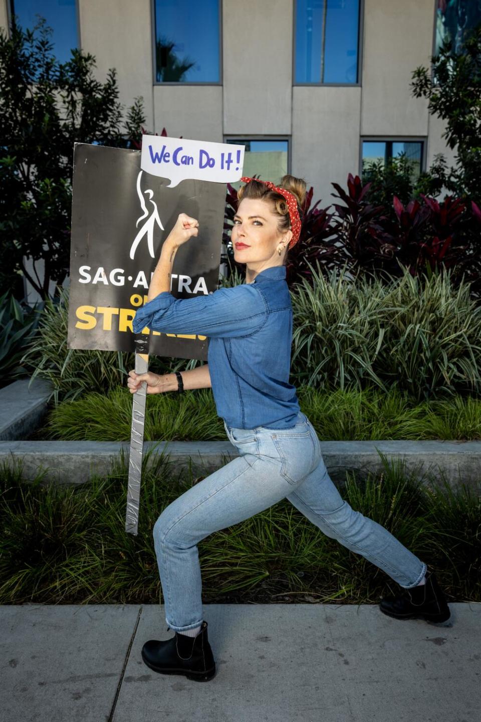 A woman dressed as Rosie the Riveter lunches forward and flexes a muscle while holding a picket sign