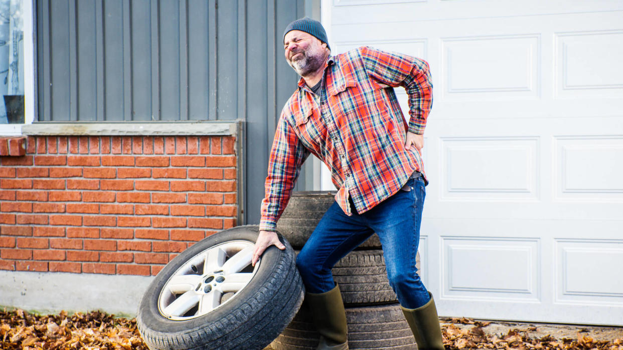 A man straining and injuring his back lifting a large truck tire improperly.