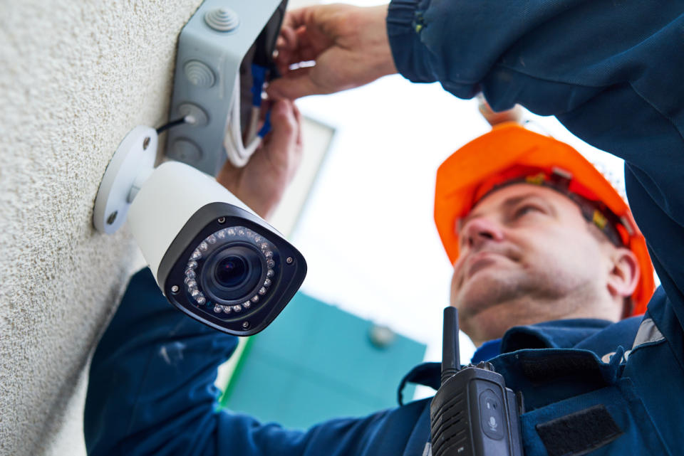 A close up of a worker in an orange hard hat installing a security camera.