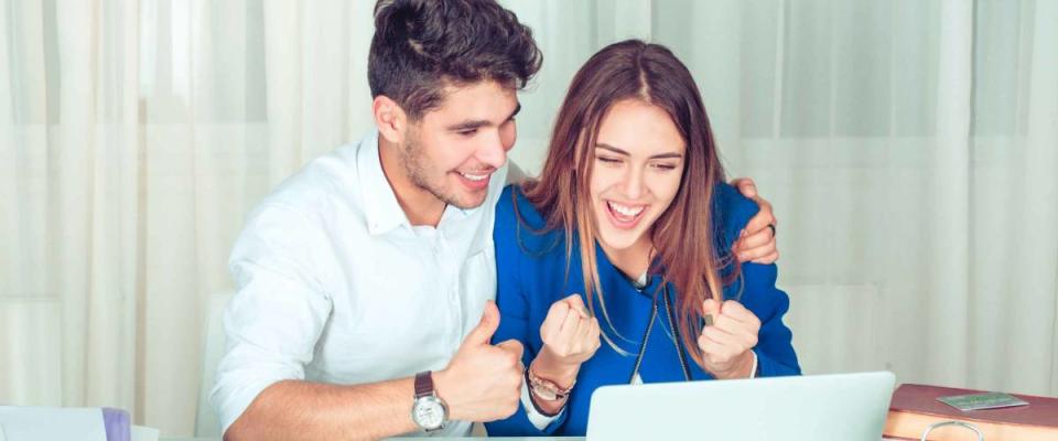 Woman sitting working at laptop in the office celebrating success pumping fists and man hugs and supports her showing thumbs up like hand gesture.