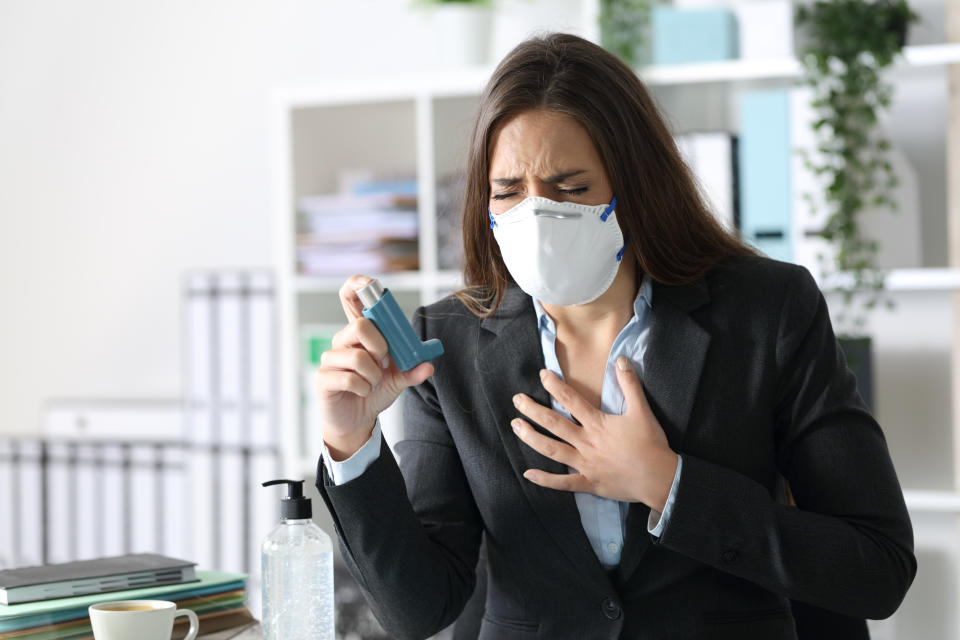 Executive woman wearing protective mask with asthma attack holding inhaler touching chest at the office