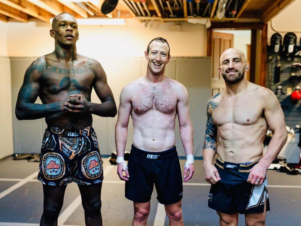 Mark Zuckerberg standing in a gym with Isreal Adesanya and Alex Volkanovski, who are all shirtless and wearing sports shorts.
