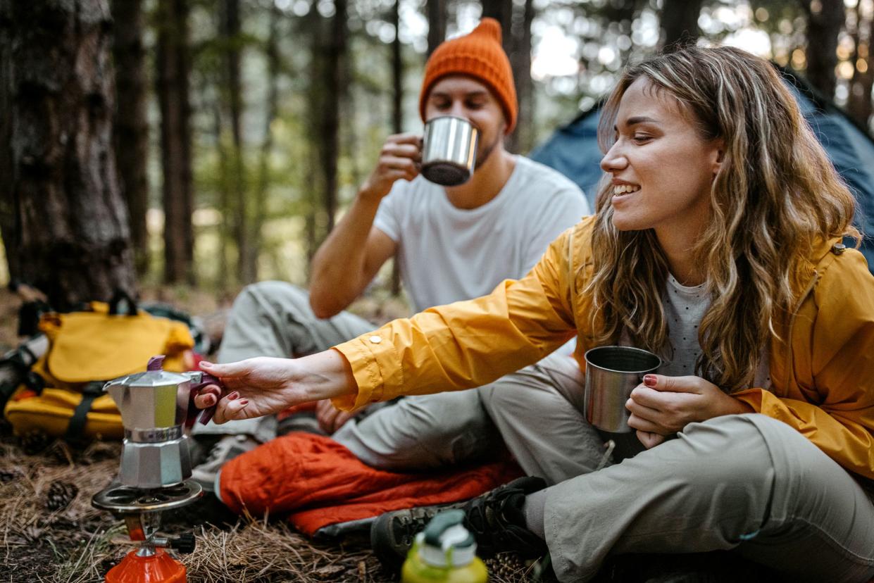 Beautiful and romantic young man and woman preparing hot coffee and tea beverage n portable stove sitting outside tent during hiking adventure