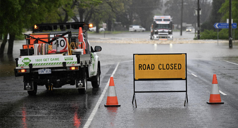 A road closed sign stands in the middle of a road while flooding can be seen in the background.