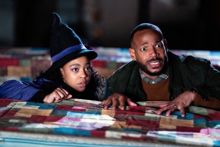 Marlon looking scared and hiding behind a table with a child who's wearing a witch's hat