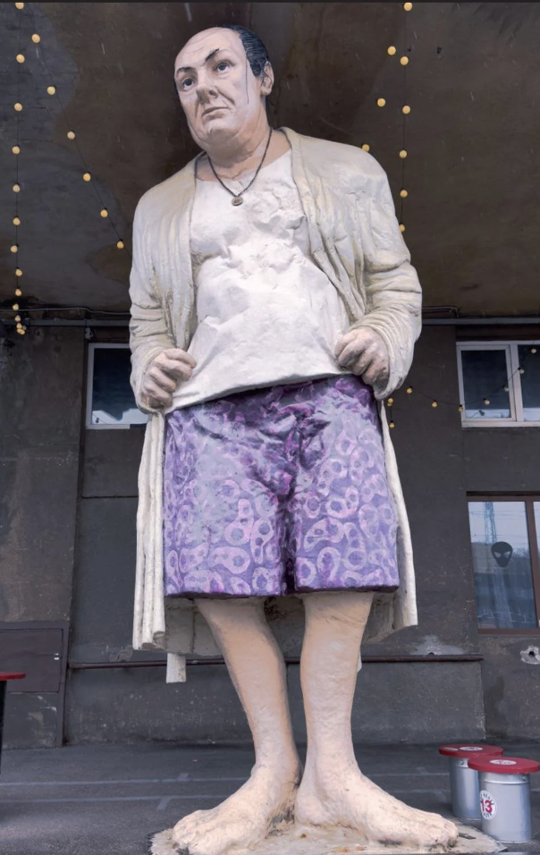 Statue of a person in a worn robe, torn shirt and patterned shorts standing barefoot. The setting appears to be an urban location with lightbulbs hanging above