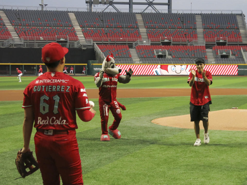 El rapero argentino Trueno, centro, aplaude después de lanzar la primera bola del partido entre los Diablos Rojos del México y el Águila de Veracruz mientras el jugador Moises Gutiérrez (61) y la mascota de los Diablos Rocco lo miran en la Ciudad de México el jueves 2 de mayo de 2024. (Foto AP/Berenice Bautista)