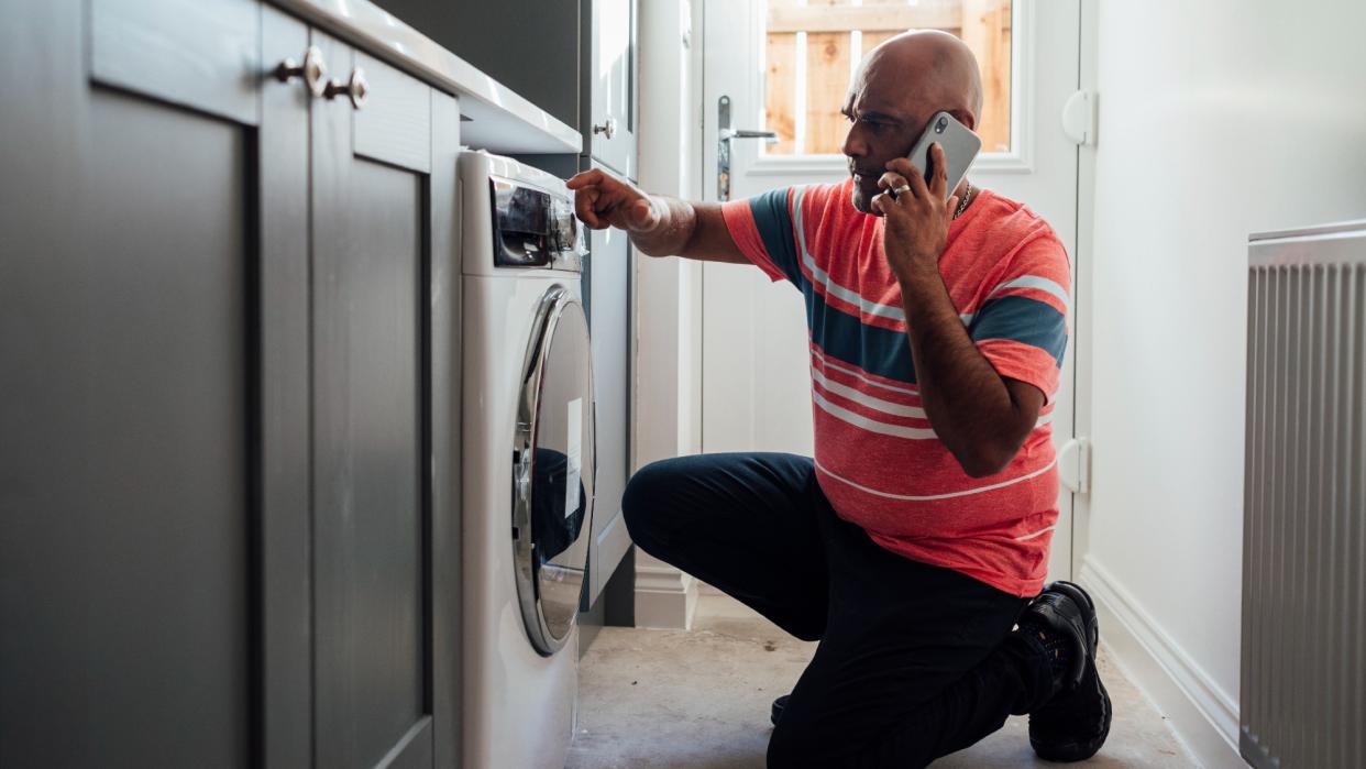  A man crouching down near his washing machine, calling an appliance repair service. 