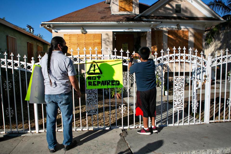 An adult and a child outside the fence of a boarded-up home.