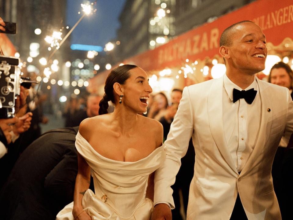 A bride and groom laugh as their wedding guests raise sparklers around them.