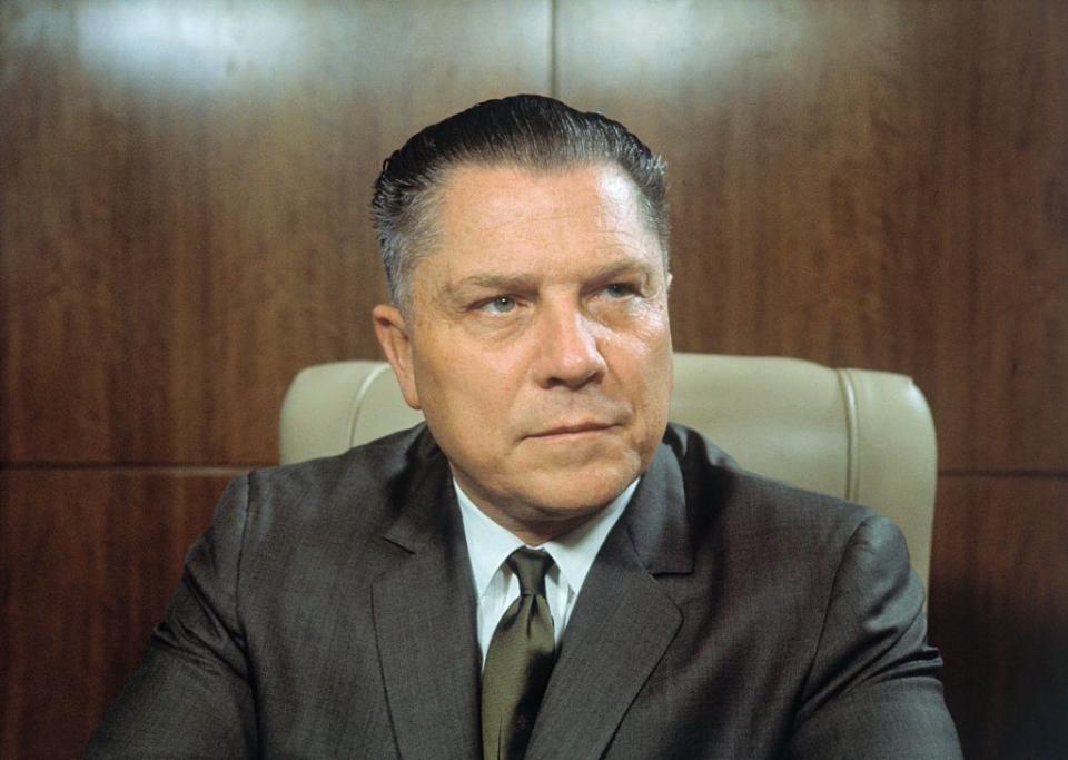 jimmy hoffa wears a suit with a tie and sits in a high backed chair in a wood paneled room, he looks to the right