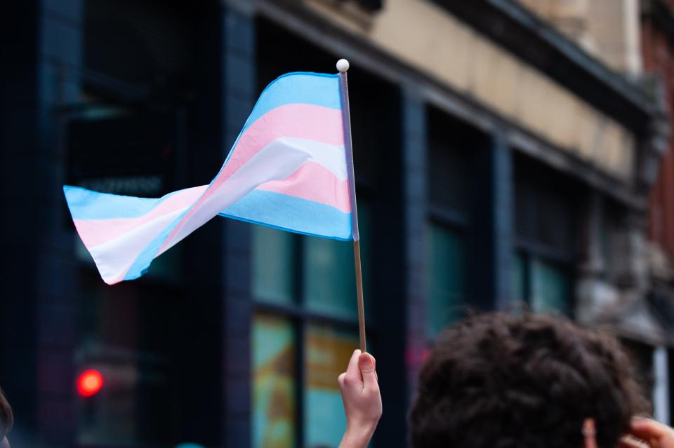 A hand holds up a small transgender pride flag in a Bristol Street. The transgender pride flag is a symbol of pride for the trans community. The blue and pink stripes represent traditional colours for a boy and girl, while the white stripe represents transitioning, intersex, neutral and undefined gender.