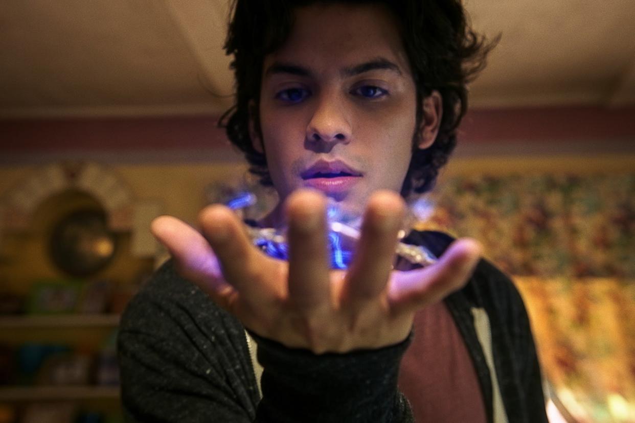 A young man in a zip-up hoodie and long, curly dark hair looks at a shining blue gizmo in his hand.