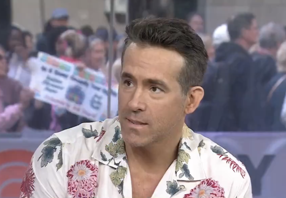 Ryan Reynolds in a floral-patterned shirt, looking off-camera with a crowd in the background