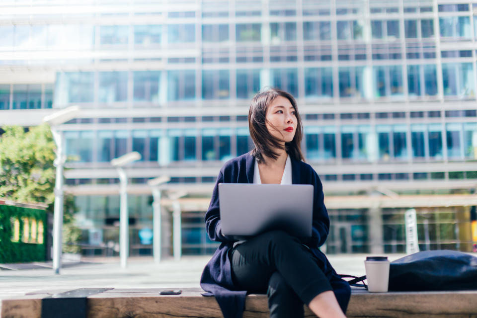 Medium shot of a motivated young Asian businesswoman working with laptop, sitting on the bench, against modern corporate buildings in the city, in daytime.