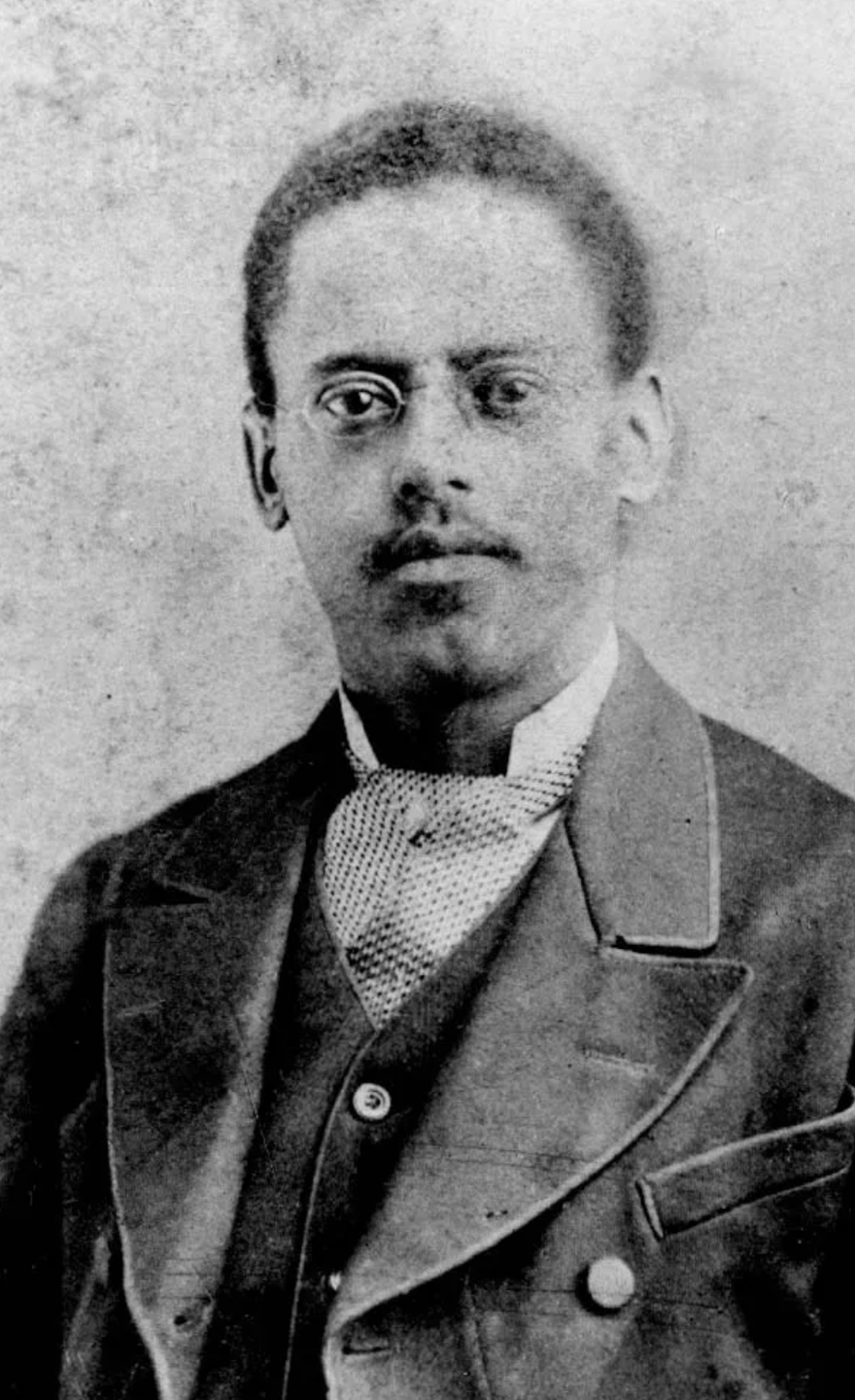 lewis howard latimer stares at the camera in a black and white photo, he wears a suit with a patterned tie and wire framed glasses