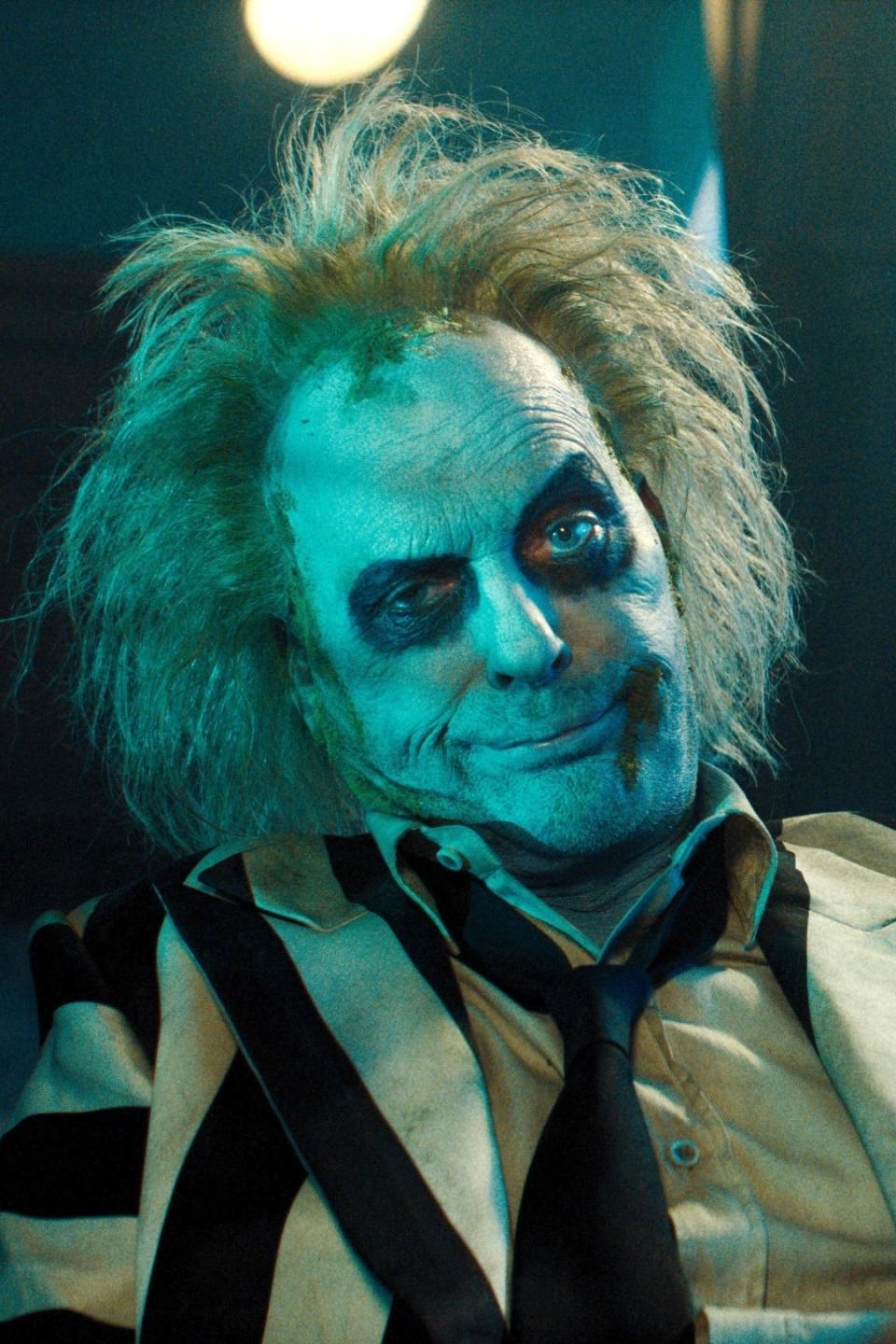 Beetlejuice, portrayed by Michael Keaton, sits with a mischievous grin in a dark room with a chaotic, disheveled appearance and a striped suit