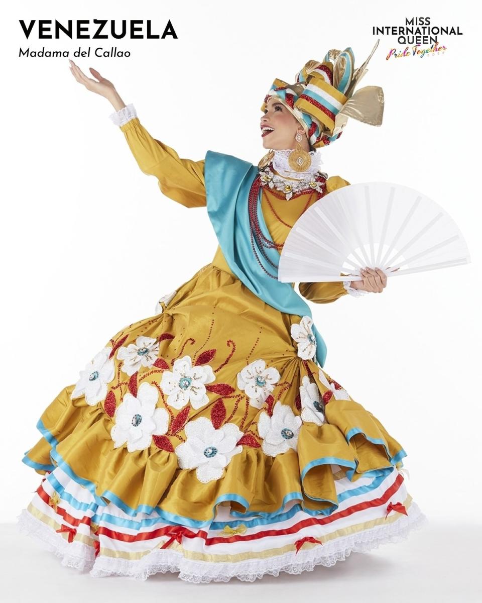 Miss Venezuela in a gold and blue dress with flowers, carrying a white fan